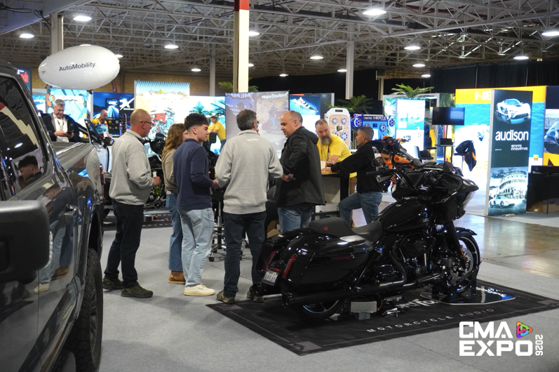 A group of people engaging in conversation at a trade show, with a black motorcycle displayed prominently in the foreground and various vendor booths visible in the background.