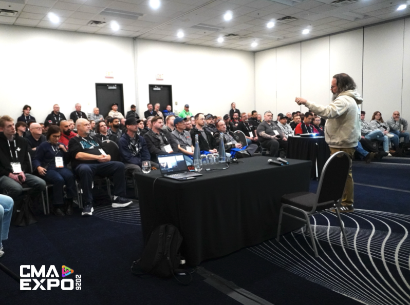 A presenter speaking to a large audience in a conference room, with attendees seated and engaged. A table in front displays equipment and water bottles.