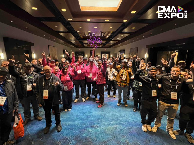 A large group of people gathered in a hallway, many wearing matching pink shirts, smiling and raising their hands in the air. The setting features a modern interior with a blue carpet and decorative lighting.