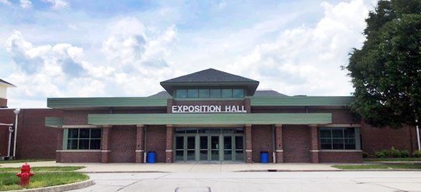 Front view of Exposition Hall, a brick building with large glass doors and a green roof, set against a partly cloudy sky.