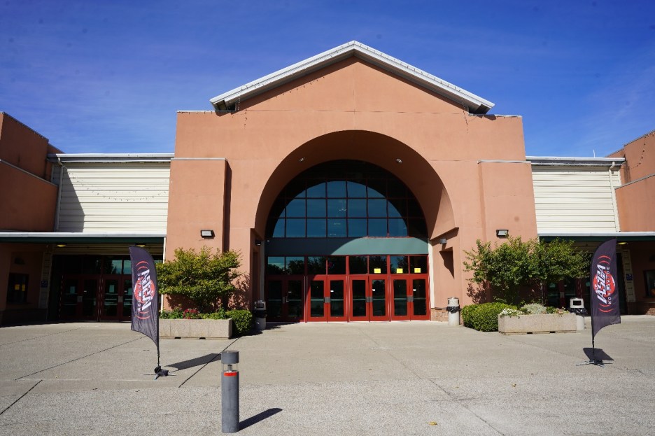 Front view of a modern building with a large arched entrance, flanked by two colorful flags and surrounded by greenery on a sunny day.