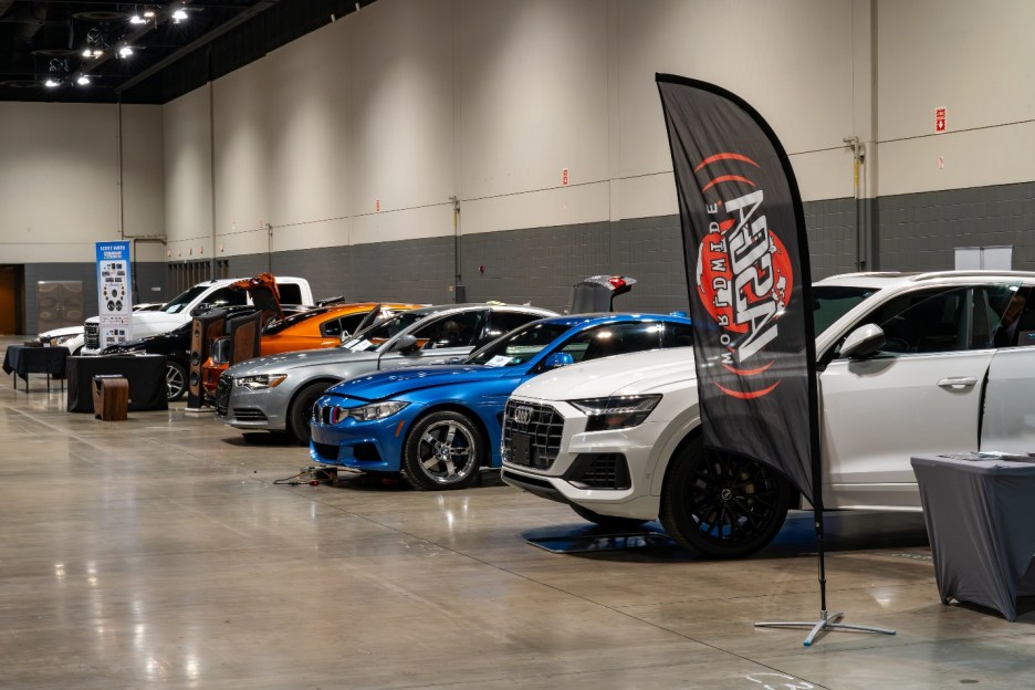 A lineup of various cars displayed at an indoor event, featuring vehicles in different colors and styles, along with a promotional banner.