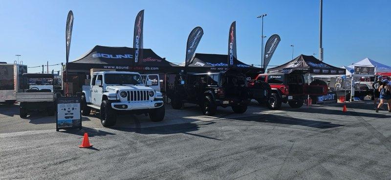A lineup of modified trucks and SUVs displayed under branded tents at an outdoor event, with clear blue skies and asphalt ground.