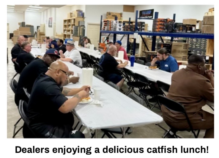 A group of individuals sitting at long tables enjoying a catfish lunch in a spacious venue.