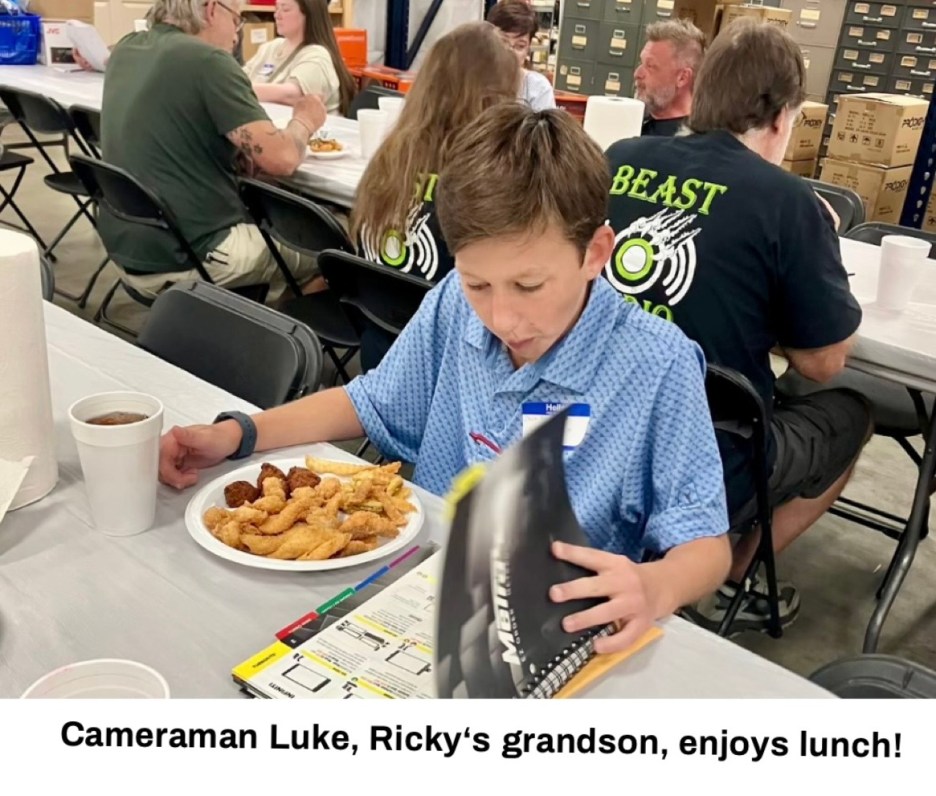 A young boy named Luke sitting at a table with a plate of food, reading a magazine, while others are seated in the background.