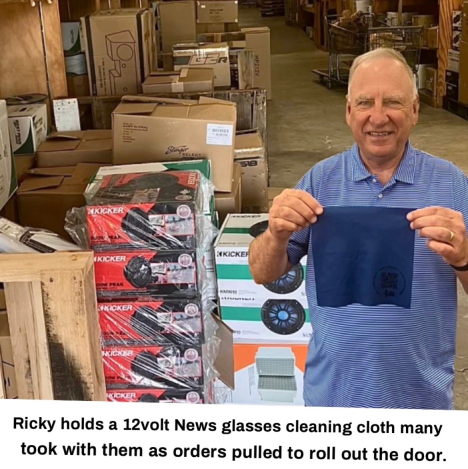 A man holding a 12volt News glasses cleaning cloth in a warehouse filled with boxes and products.