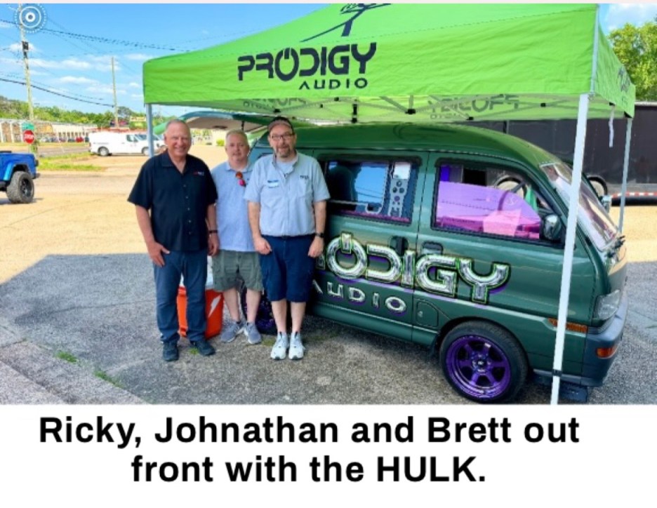 Three men stand in front of a green van with purple rims, under a green tent displaying 'PRODIGY AUDIO'.