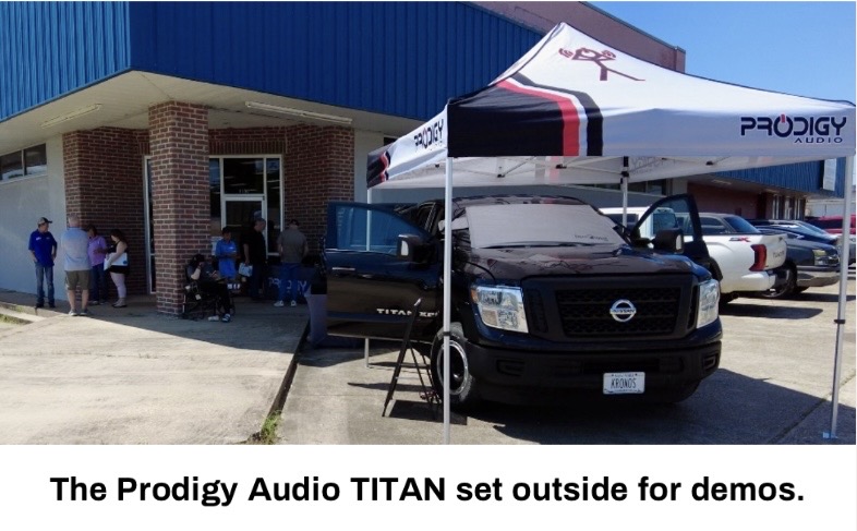 Prodigy Audio TITAN vehicle set up outside a building under a branded tent, with people gathered around for demonstrations.