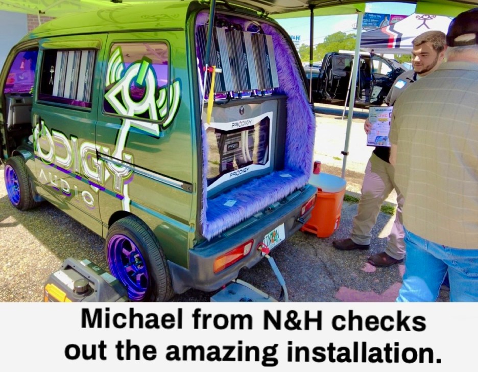 A customized van showcasing impressive audio equipment installation, featuring vibrant decor and purple accents, with two individuals examining the setup.