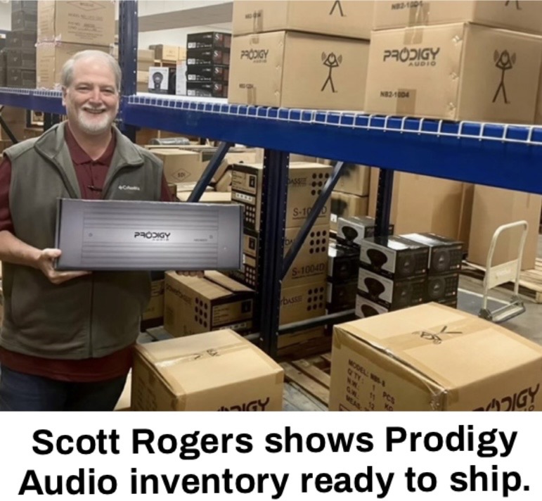 A man holding a Prodigy Audio product stands in a warehouse filled with boxes of audio equipment and inventory.