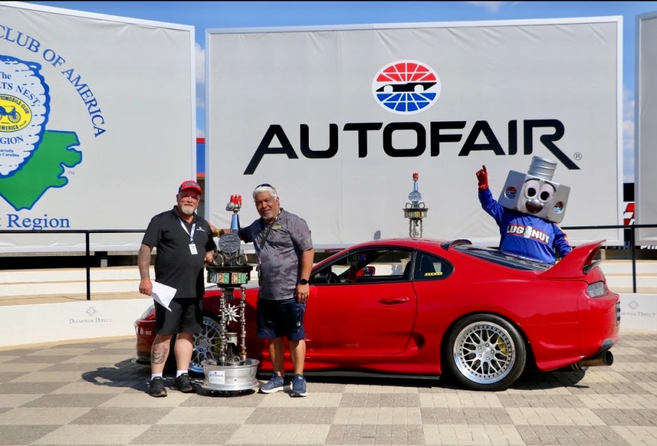 Two men pose with a trophy and a humorous mascot beside a red sports car at an automotive event, with large banners in the background.