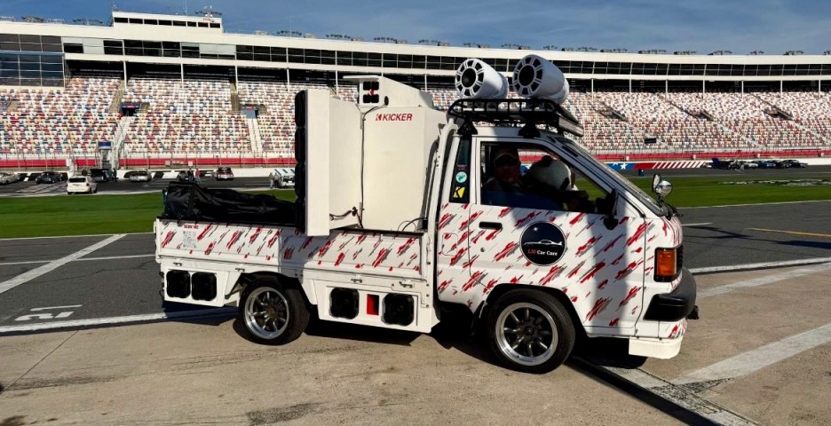A customized small truck decorated with bright graphics, equipped with large speakers and a sound system, parked on a racetrack with stadium seating in the background.