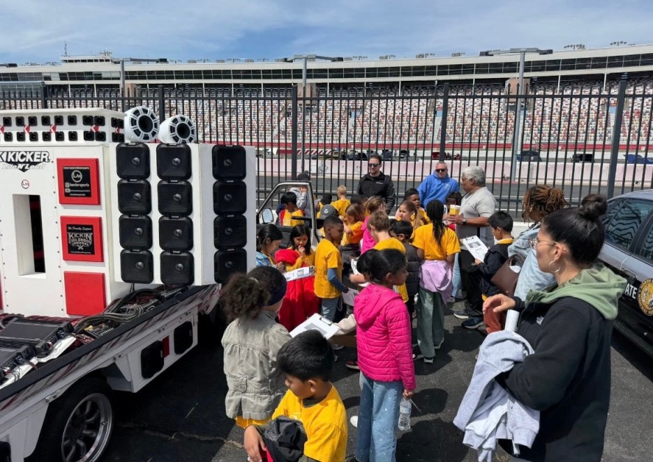 A group of children in yellow shirts gathered around a large sound system on wheels, with adults supervising them at a racetrack. The background features a grandstand and fencing.