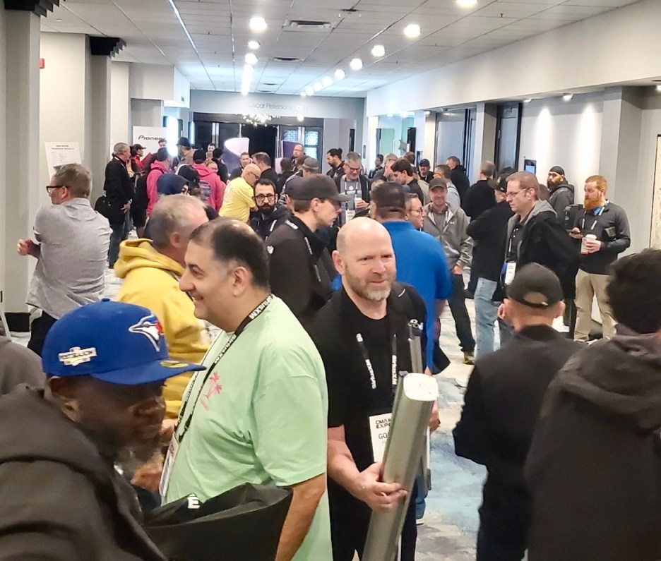 A crowded hallway filled with people attending an event, some engaging in conversation while others are walking. Various individuals are holding drinks and items, with colorful clothing and name tags visible.