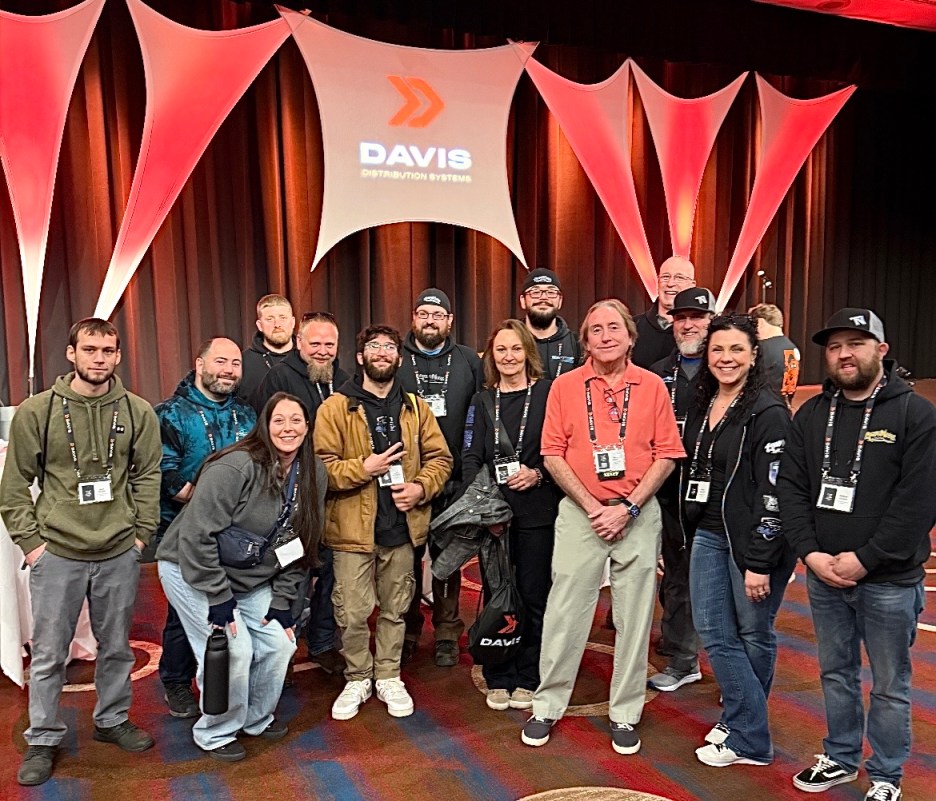 A group photo of attendees posing together in front of a stage with a 'Davis Distribution Systems' banner, featuring a diverse group of men and women, smiling and wearing name badges.