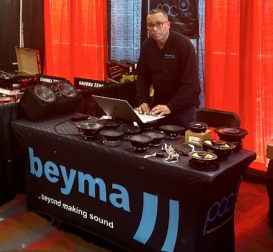 A vendor at a trade show, standing behind a table displaying various audio equipment and speakers, with a laptop open in front of him and a backdrop of red curtains.