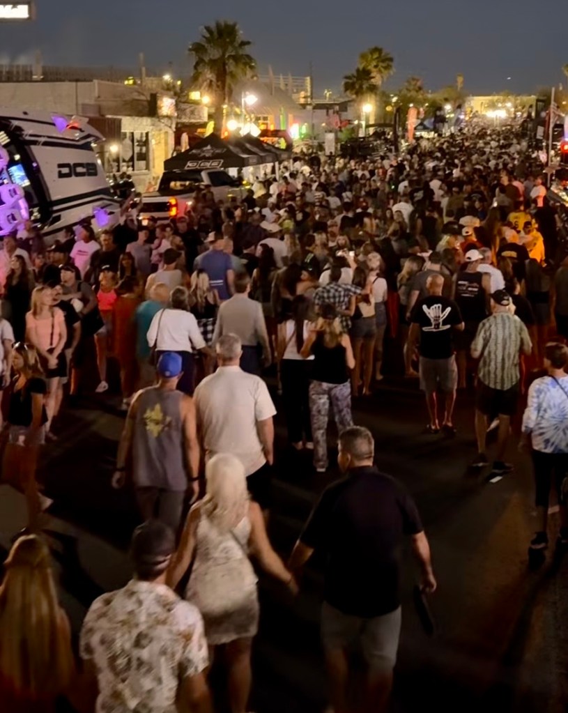 A large crowd of people walking along a street during the night, with palm trees and colorful lights in the background.