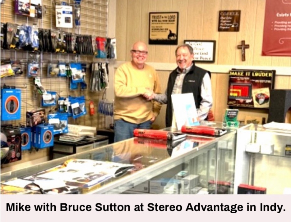 Two men shaking hands inside a retail store filled with audio equipment and accessories. The man on the left is wearing a yellow sweater, while the man on the right is in a black vest. Various products and displays are visible in the background.