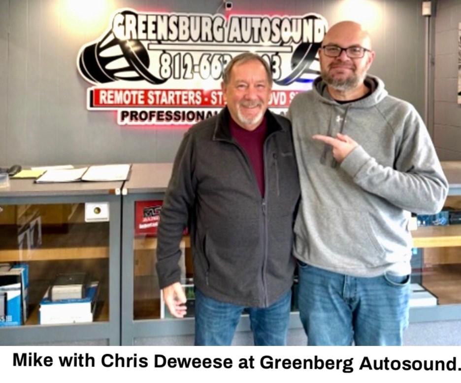 Two men posing together at Greensburg Autosound, with a sign in the background displaying the business name and services offered.
