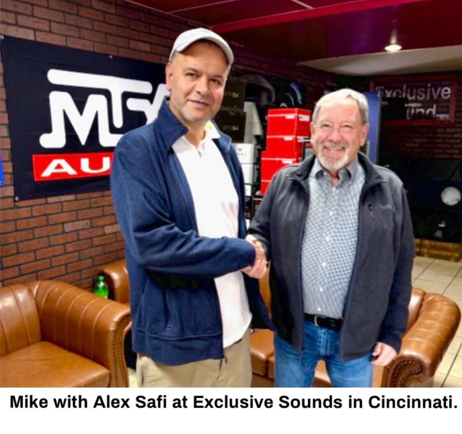 Two men shaking hands inside a store, with a brown sofa in the foreground and sound equipment in the background. A banner with 'Exclusive Sounds' is visible.