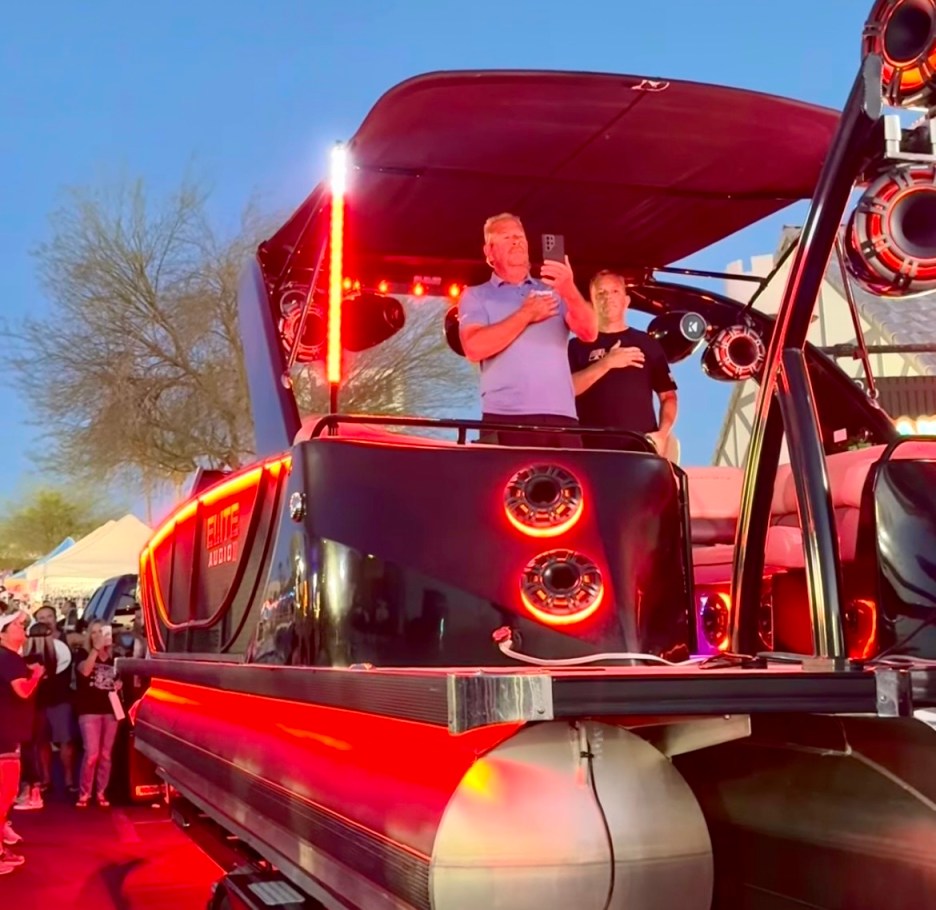 A group of men on a stylish black boat with red LED lights, set against a dusk background. One man is holding a phone while another stands beside him.