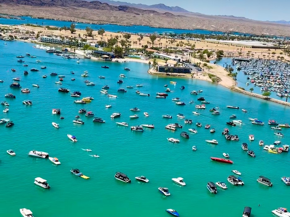 Aerial view of a vibrant blue lake filled with numerous boats and recreational watercraft, surrounded by sandy beaches and palm trees.