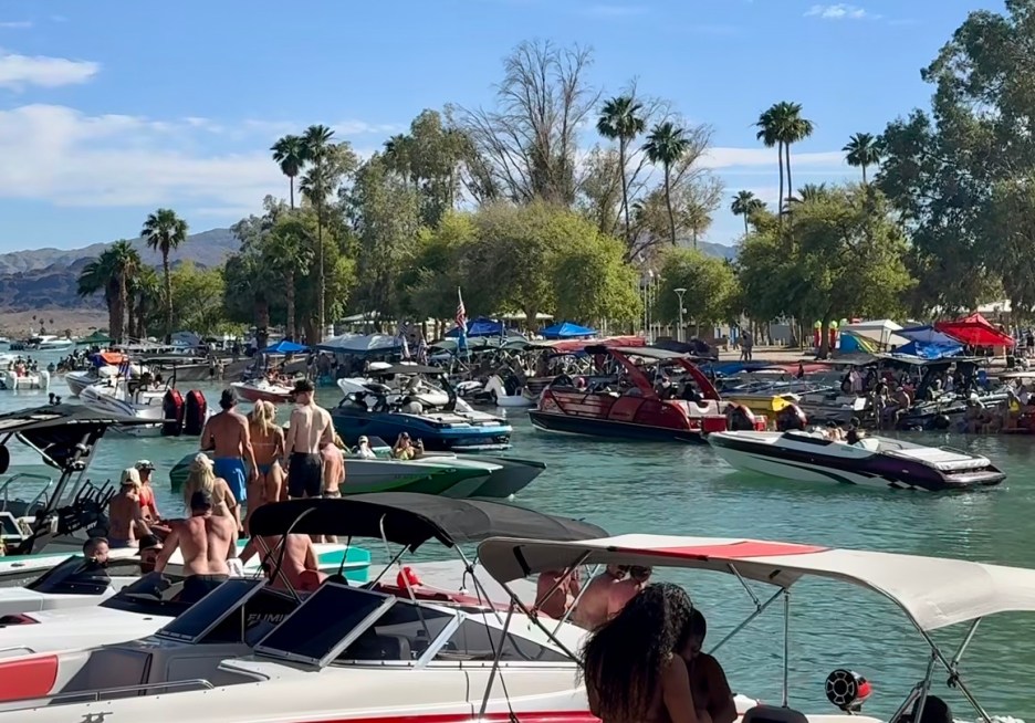 A busy river scene with multiple boats and people enjoying a sunny day. Palm trees line the shore, and several tents can be seen on the banks.