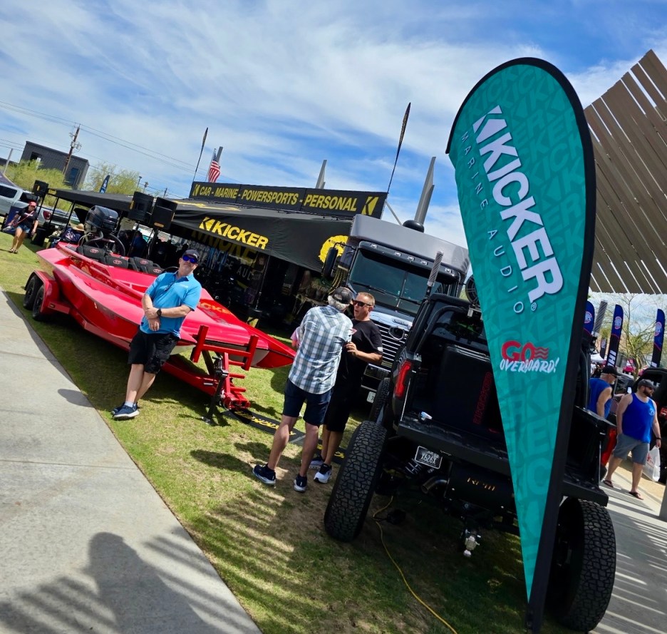 Outdoor event with a red boat and a black truck, featuring Kicker branding. People are interacting near the display, with banners in the background.