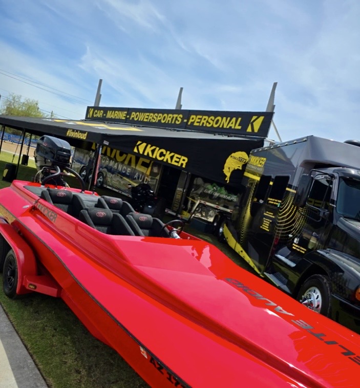 A bright red boat displayed in front of a KICKER performance audio booth with promotional signage, surrounded by vehicles and under a clear blue sky.