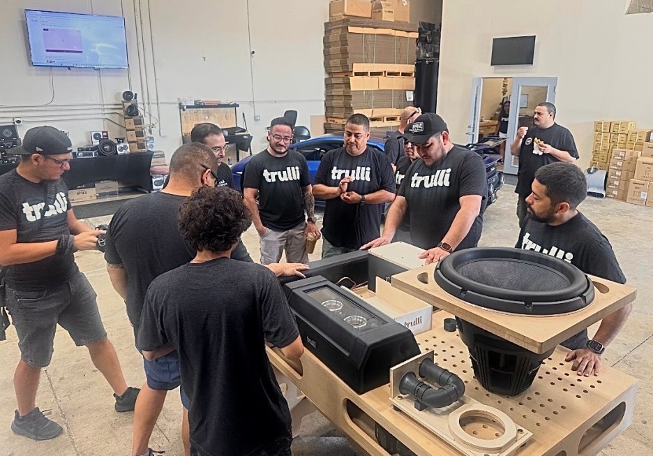 A group of eight individuals gathered around a table examining audio equipment components, all wearing black shirts with the logo 'trulli'. The setting is in a workshop with various audio equipment in the background.