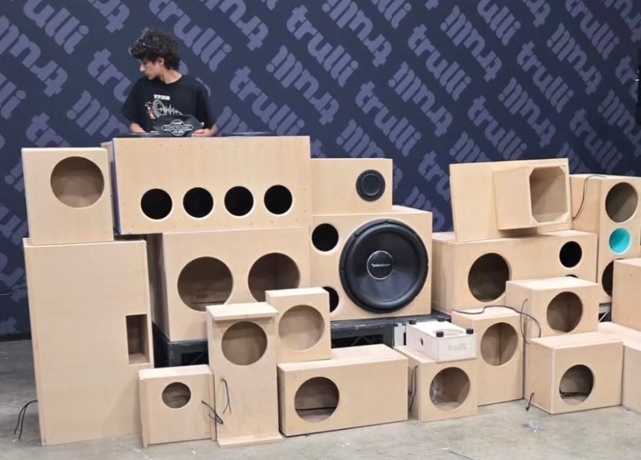 A young person standing beside a display of wooden speaker boxes in various sizes and shapes, with a large circular speaker prominently featured.