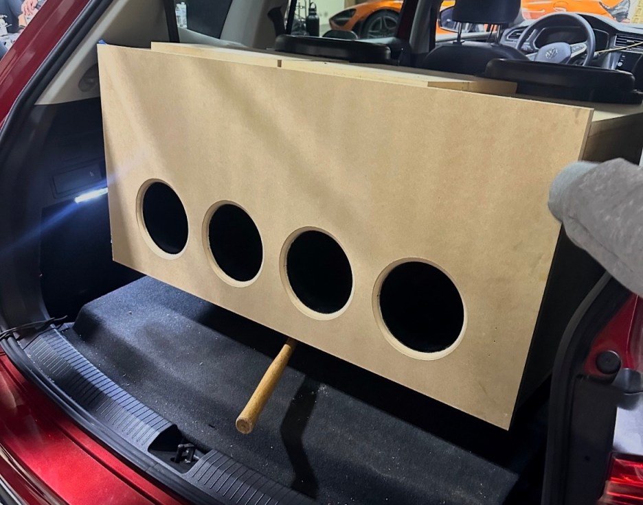 A wooden speaker box with four circular openings is being held in the trunk of a car.