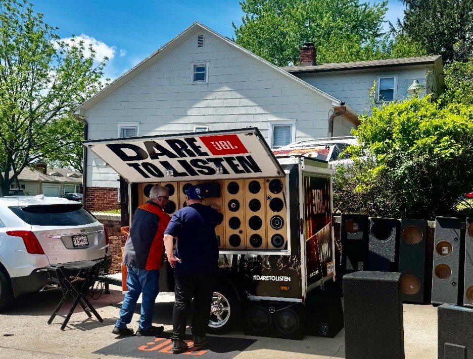 A trailer displaying various speakers and audio equipment, with two people interacting in front. The trailer has a sign that reads 'DARE TO LISTEN' and features the JBL logo. In the background, there is a house and a parked white vehicle.