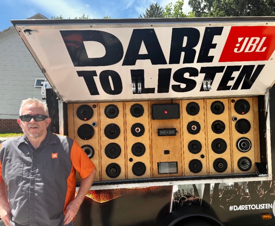 A man wearing sunglasses stands next to a large speaker display trailer featuring multiple speakers, with the words 'DARE TO LISTEN' prominently displayed above.