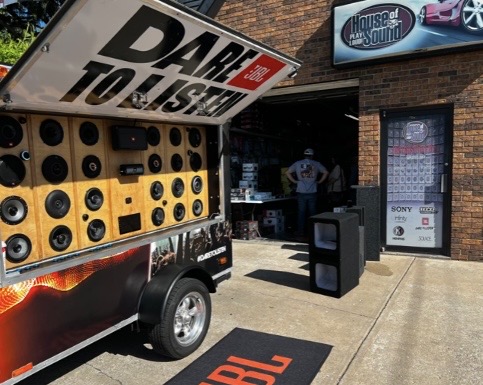 A mobile sound system trailer displaying numerous speakers with the slogan 'DARE TO LISTEN,' positioned outside a store called 'House of Sound.'