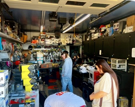 A cluttered garage filled with various electronic products, including audio equipment and accessories, with people browsing and organizing items.