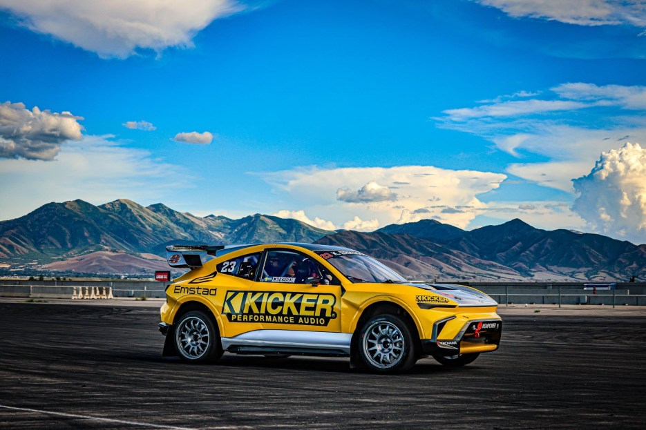 A yellow and black racing car with the Kicker Performance Audio logo parked on a racetrack, set against a backdrop of mountains and a blue sky with clouds.