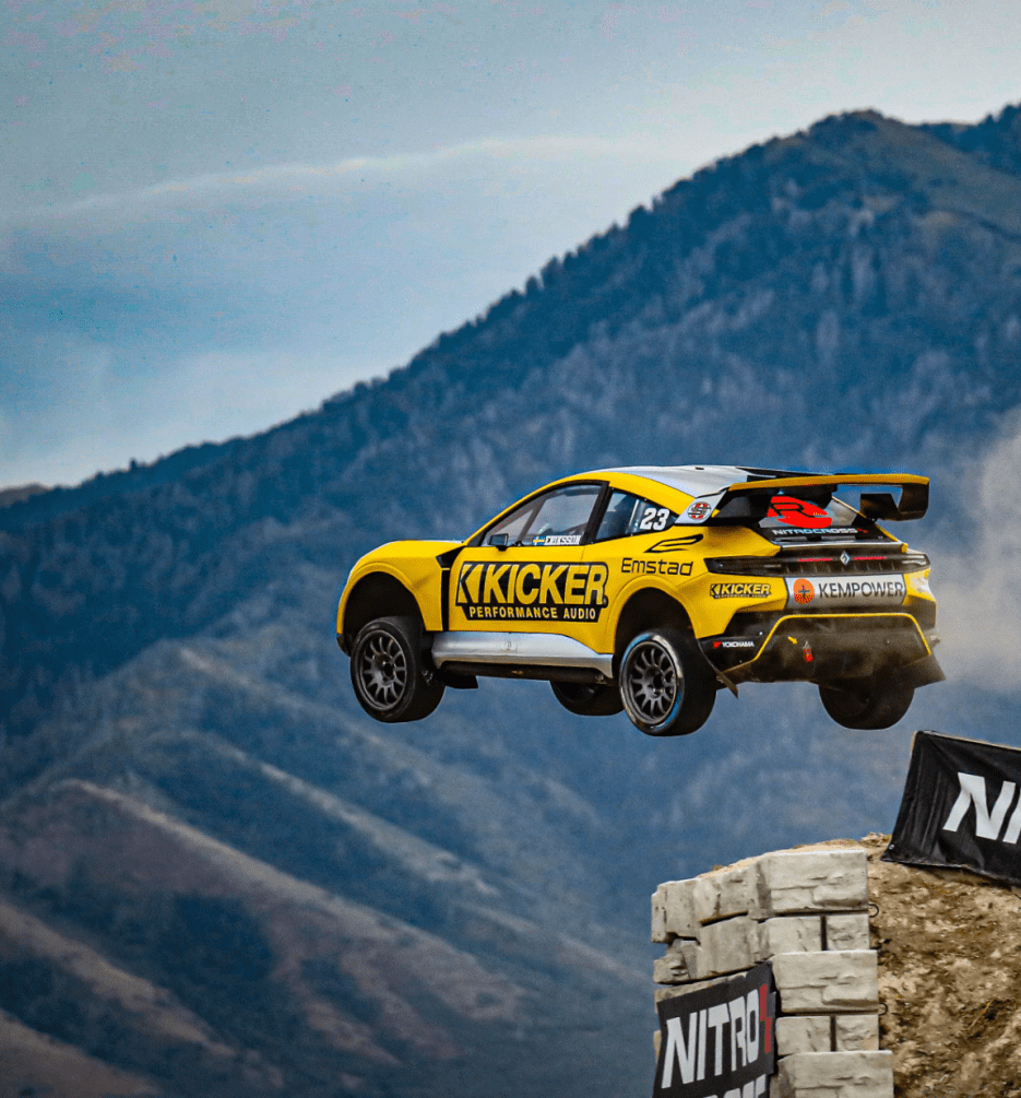 A yellow rally car jumps off a ramp with mountains in the background, showcasing extreme motorsport action.