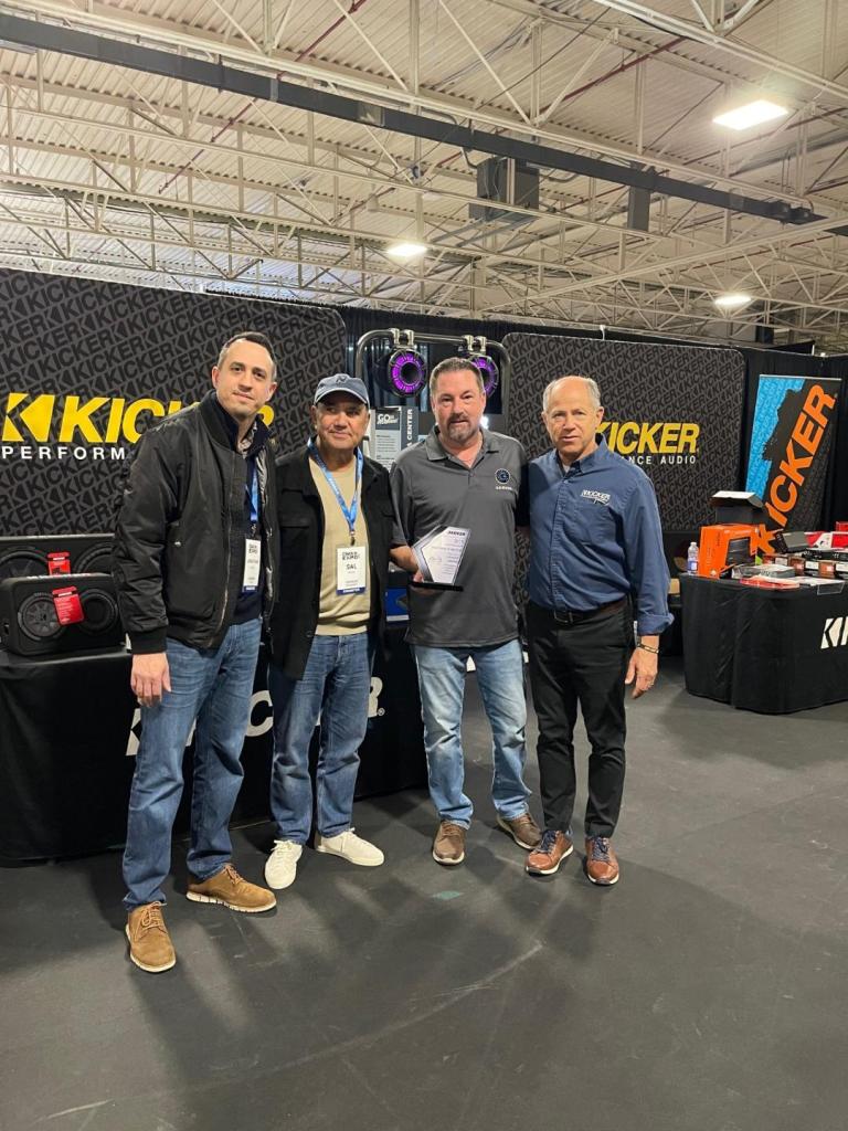 Four men posing together at a KICKER audio booth at an event, showcasing audio equipment in the background.