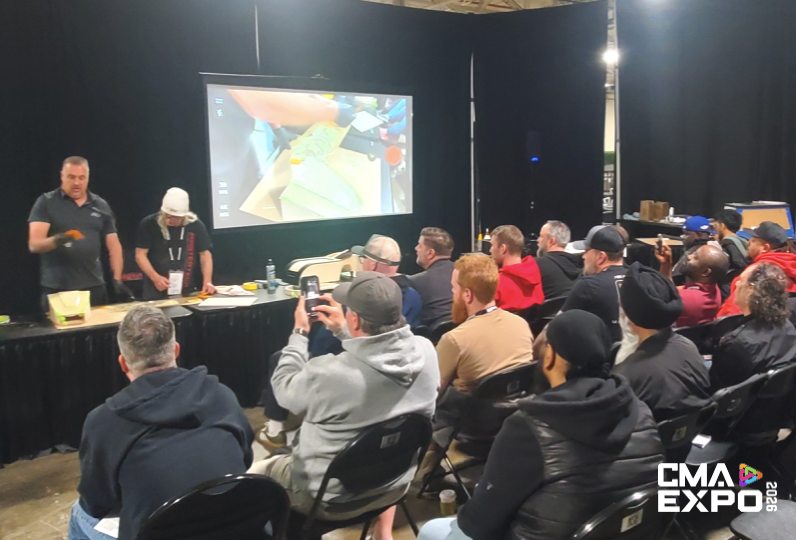 A group of attendees watching a presentation at CMA Expo, with two speakers demonstrating techniques on stage and a large screen displaying close-up visuals.