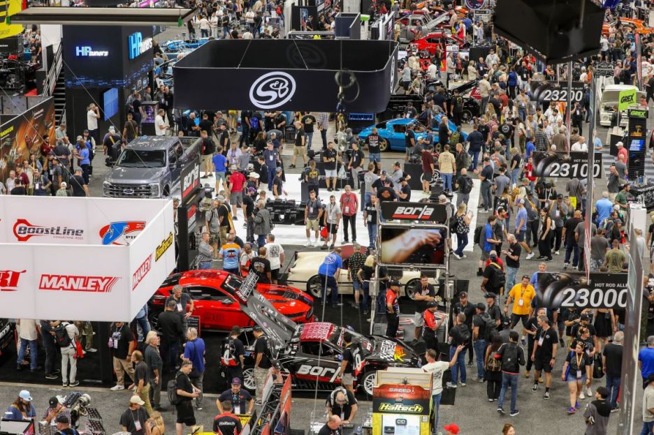 Aerial view of a crowded automotive trade show with numerous booths and displays, showcasing cars and automotive parts, with attendees browsing around.