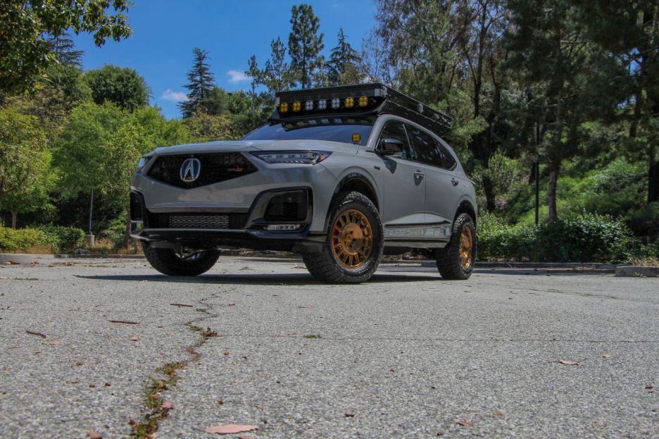 A stylish gray SUV with a distinctive front design, equipped with off-road wheels and a roof rack featuring multiple lights, parked in a natural outdoor setting.
