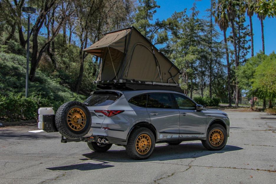 A gray SUV equipped with a rooftop tent parked in a sunny outdoor setting, surrounded by trees.