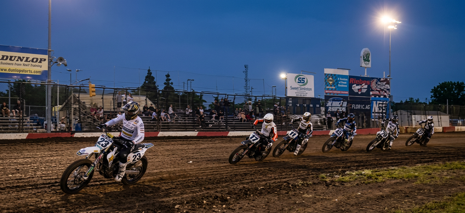 A group of motorcycle racers competing on a dirt track during dusk, with spectators in the background and advertising banners along the track.