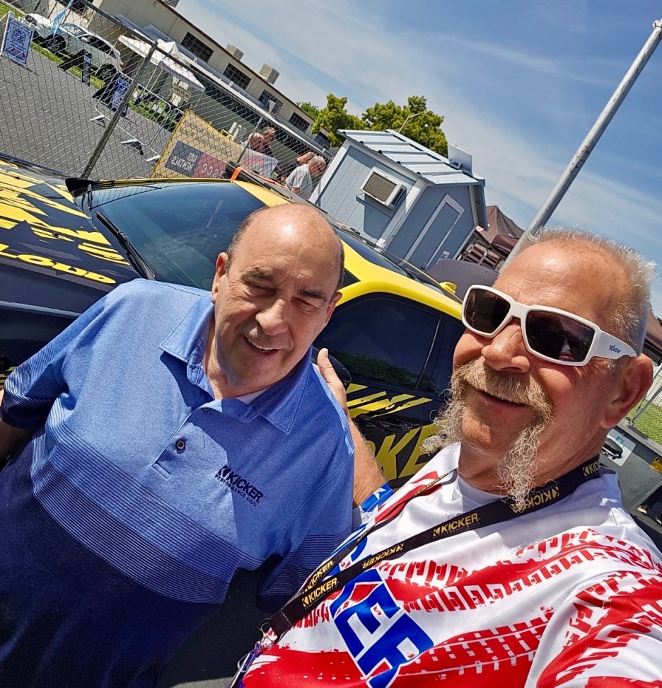 Two men posing for a selfie in front of a car with Kicker branding. One man is wearing sunglasses and a patterned shirt, while the other is wearing a blue polo shirt. The background shows a clear sky and event setup.