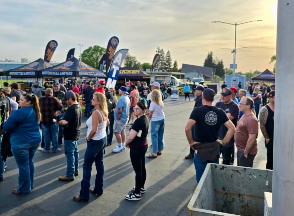 A crowd of people lined up at an outdoor event with various tents displaying logos, including 'Motorcycle Lawyers' and 'Kicker,' set against a sunset sky.