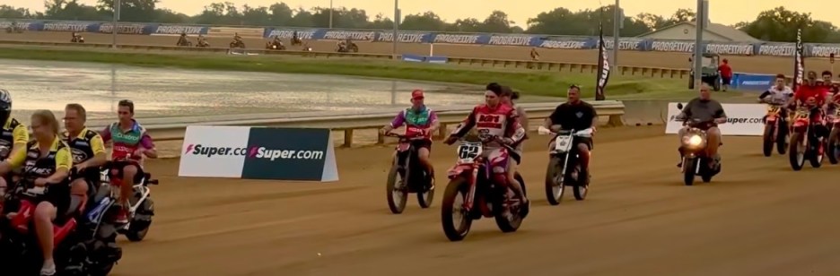 A group of motorcycle riders lined up on a dirt track, with some standing and others seated. Colorful banners and a scenic background featuring a body of water are visible, indicating a racing or motorsport event.
