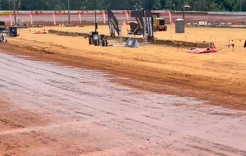 View of a dirt racing track with wet soil and several banners, creating a pre-race atmosphere at an outdoor event.