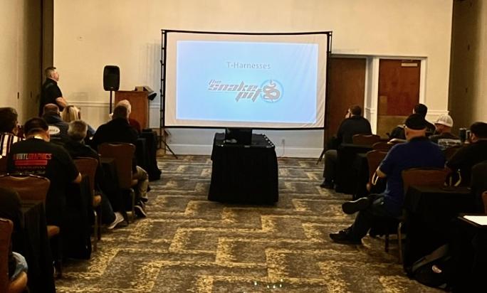 A conference room with an audience seated at tables, listening to a speaker presenting on T-Harnesses, displayed on a screen with a logo.