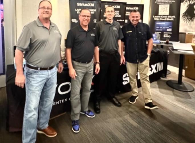 Four men standing together in front of a SiriusXM promotional booth, wearing matching gray shirts and smiling at the camera.
